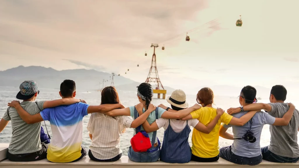 Un gruppo di sette amici seduti su una panchina, abbracciati, guardando il mare con montagne e funivie sullo sfondo.